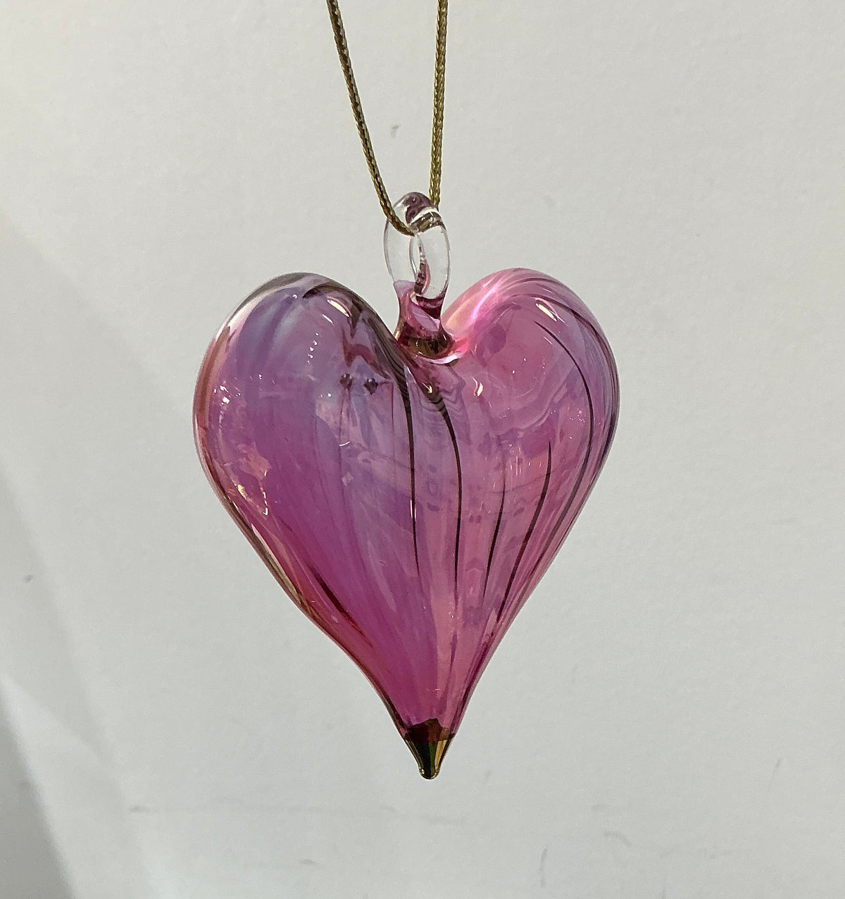 A pink glass heart-shaped pendant necklace hanging from a gold chain against a white wall, with a white radiator and household items visible below.
